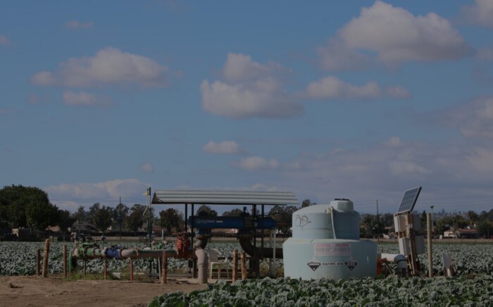 Farm irrigation equipment, including pipes, a filtration unit under a metal canopy, and a chemical injection tank, standing in a field of leafy green crops in Ventura County with houses in the distance.