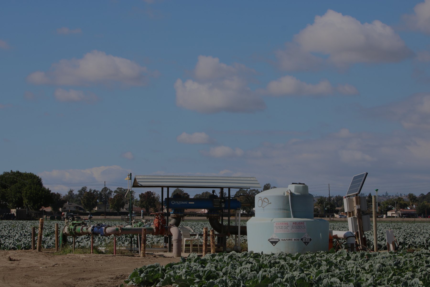 Farm irrigation equipment, including pipes, a filtration unit under a metal canopy, and a chemical injection tank, standing in a field of leafy green crops in Ventura County with houses in the distance.